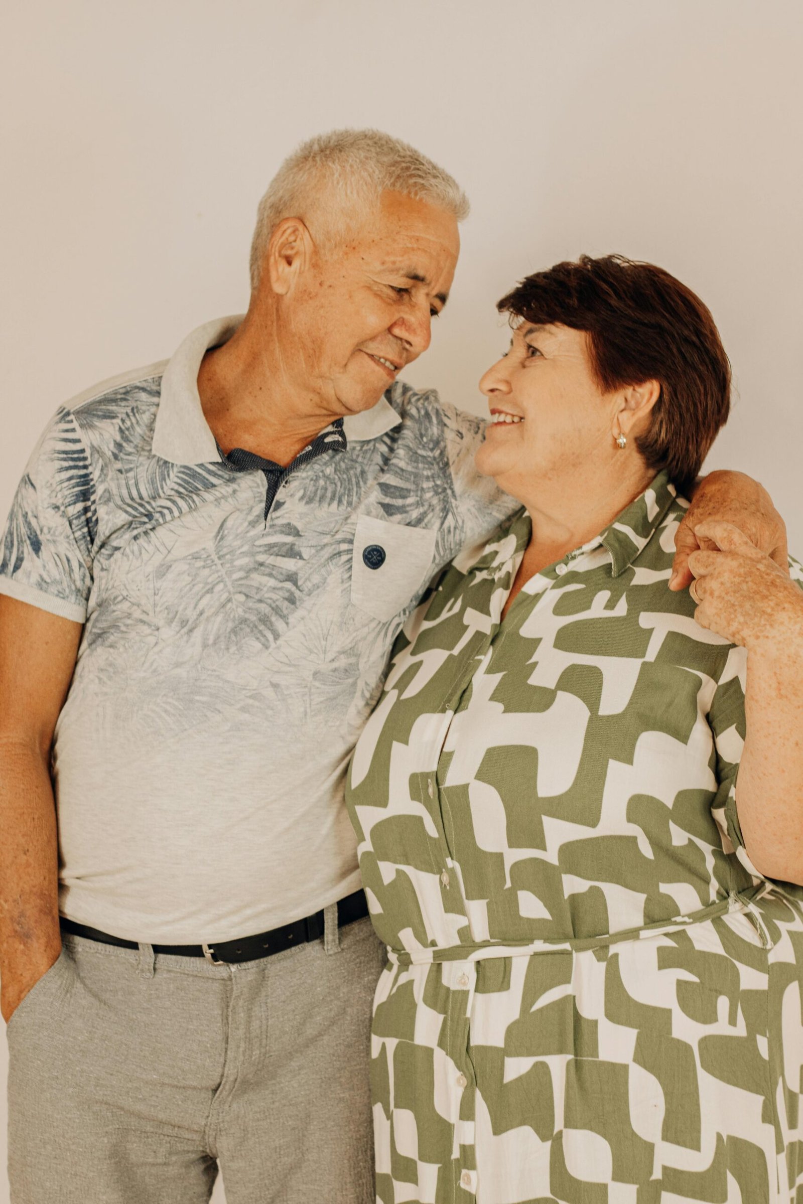 Elderly couple standing face to face, sharing a loving embrace with a warm smile in a studio setting.